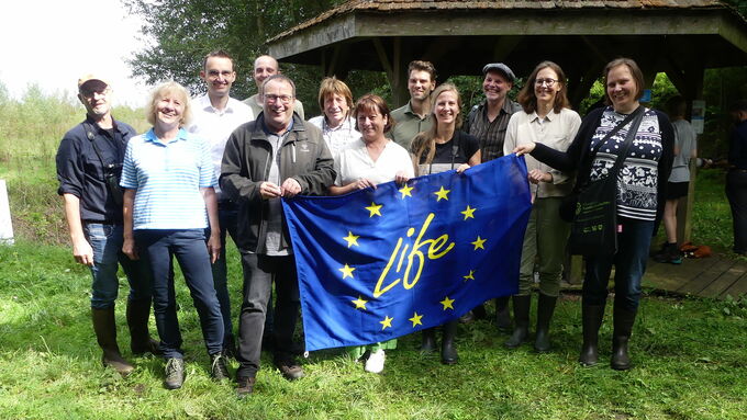 Zum Gruppenfoto mit der LIFE-Fahne versammelten sich im Oppenweher Moor am Info-Pavillon (von links nach rechts) Peter Germer (Leiter der BUND- Station Diepholzer Moorniederung), Cornelia Schmelzer (Fraktionsvorsitzende der Grünen), Frank Rabe (Stellvertret. CDU-Fraktionsvorsitzender), Alexander Menge (BUND Diepholzer Moorniederung), Umweltminister Oliver Krischer, Alfred Borgmann (CDU-Fraktionsmitglied), Bianca Winkelmann (MdL), Sönke Tielbürger (Leiter der Unteren Naturschutzbehörde Minden-Lübbecke), Luisa Stemmler (BUND Diepholzer Moorniederung), Rainer Gunia (Untere Naturschutzbehörde Minden-Lübbecke, Martina Vortherms (Leiterin Umweltamt Kreis Minden-Lübbecke) und Dr. Martina Raffel (IP-LIFE, Bezirksregierung Münster).