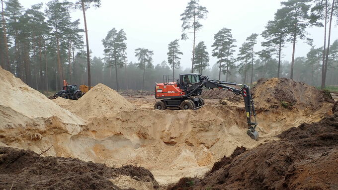 Ein Bagger gräbt eine Vertiefung in den Sandboden und lagert den Sand seitlich zwischen.